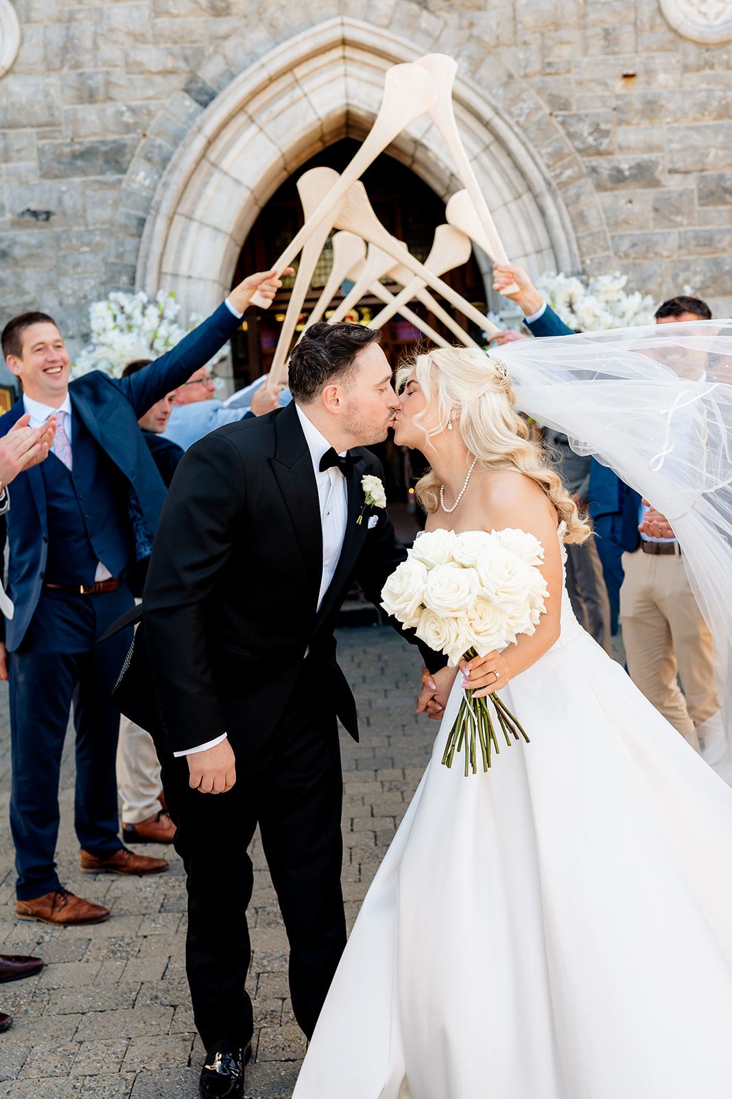 Wedding celebration with guests holding oars in arch formation outside stone church as couple shares romantic moment.