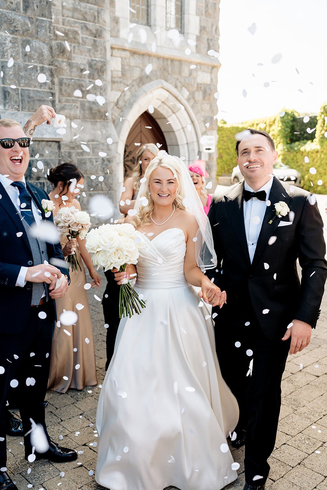 Wedding party exits church as confetti rains down on their celebration with stone archway in background.