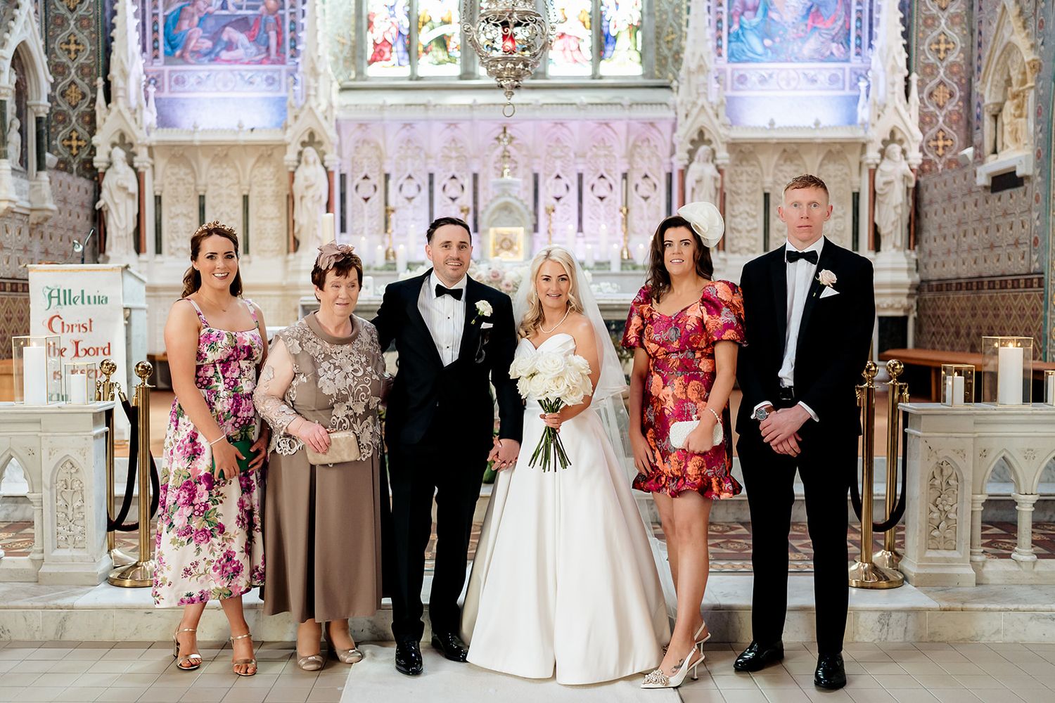 Wedding party stands together in an ornate church sanctuary with stained glass windows and decorative altar.