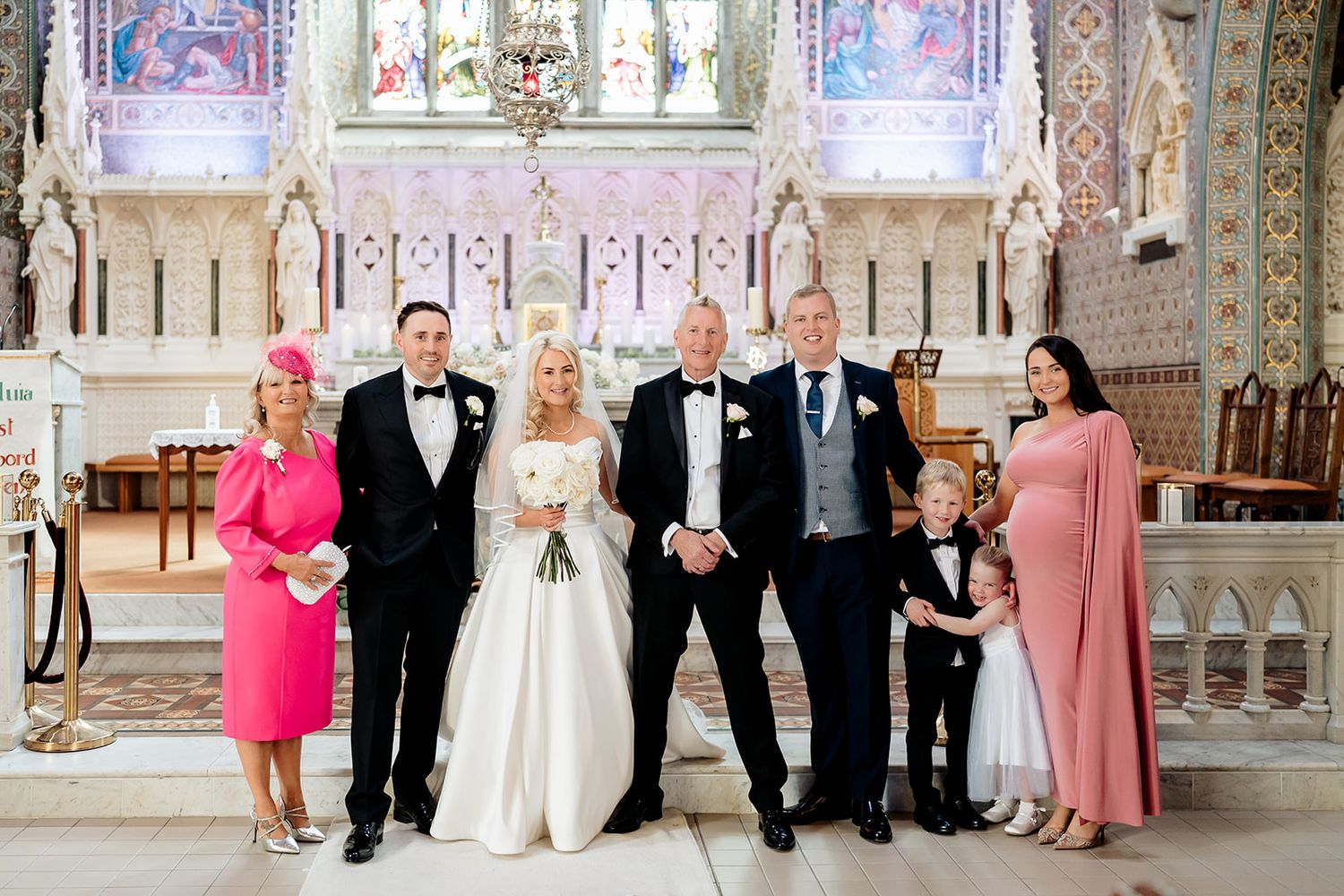 A group poses together in formal attire inside an ornate church with stained glass windows and decorative architecture.