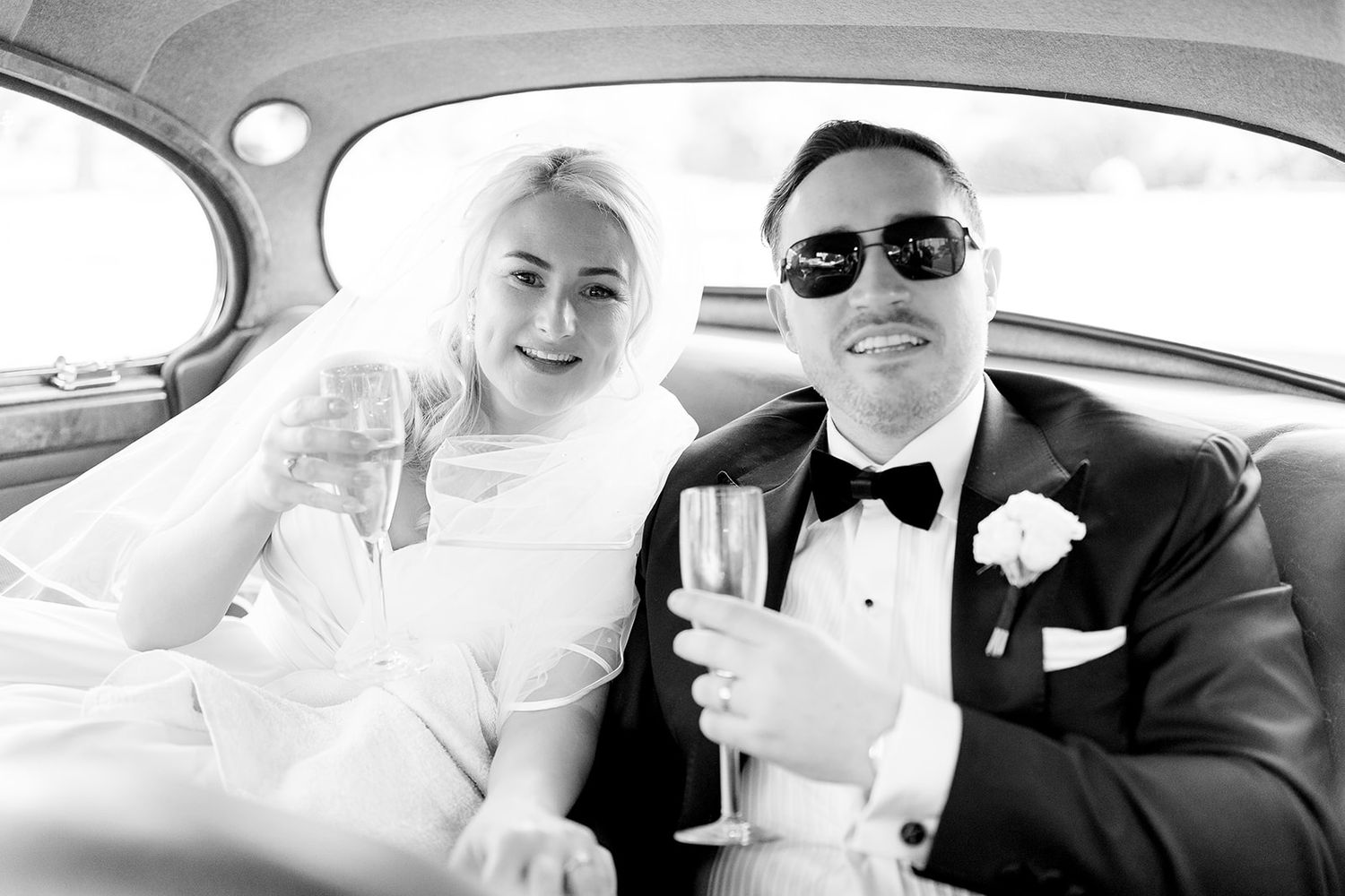 Black and white photo of newlyweds celebrating with champagne while seated in a classic car.