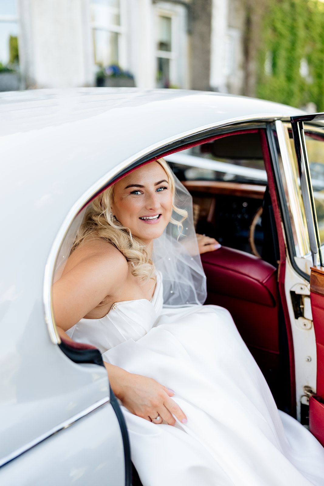A bride sits smiling in a classic white vintage car wearing a white wedding gown and veil.