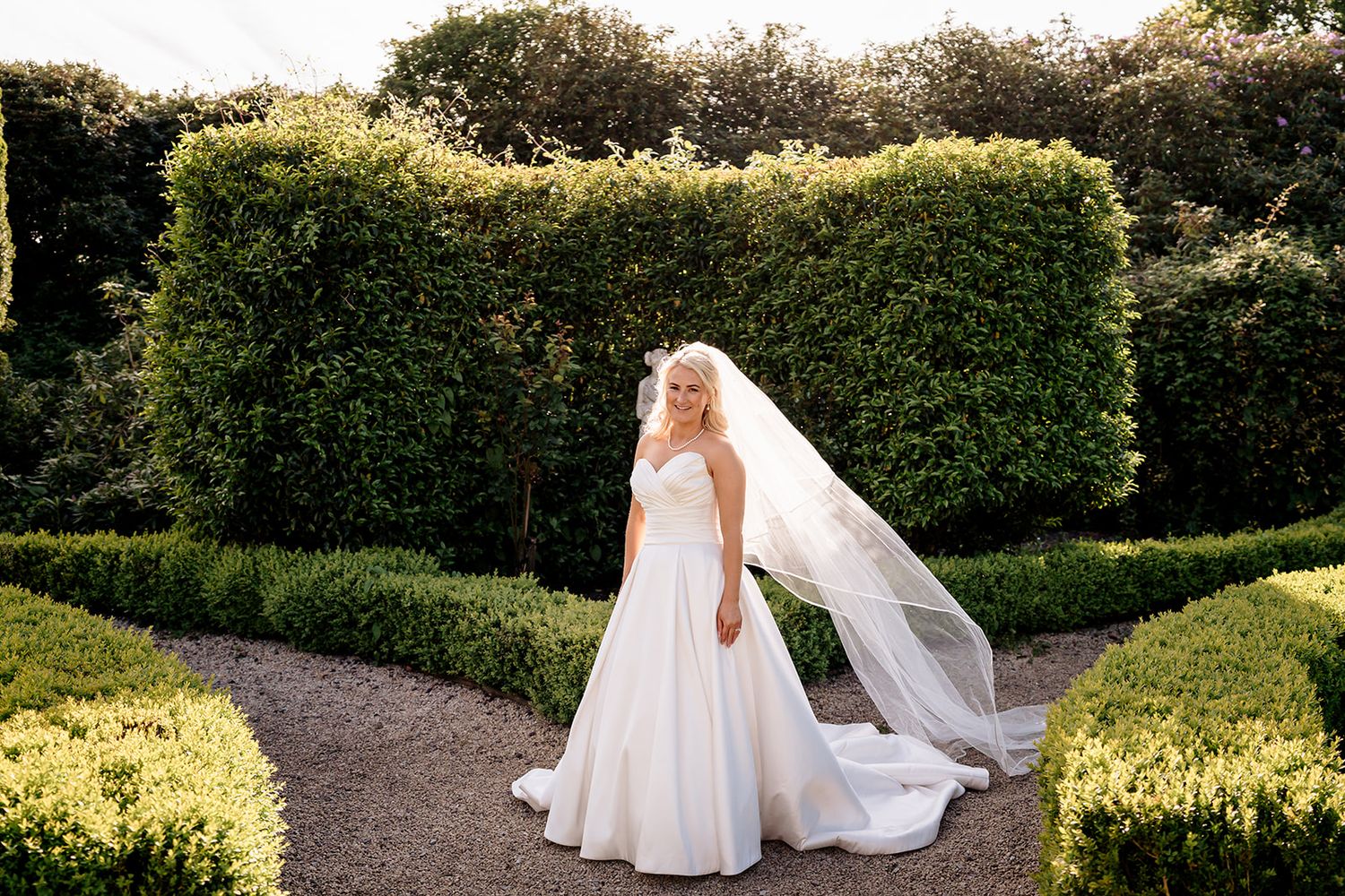 A bride in a flowing white gown and long veil stands on a garden path surrounded by manicured hedges on a sunny day.