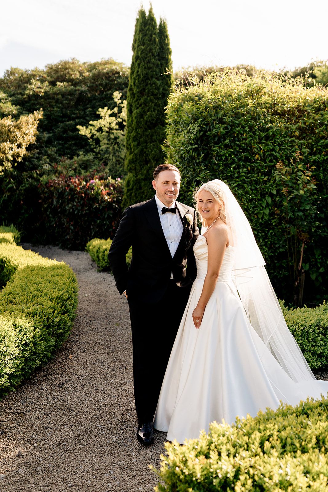 A newlywed couple walks along a garden path surrounded by manicured hedges and cypress trees on a sunny day.