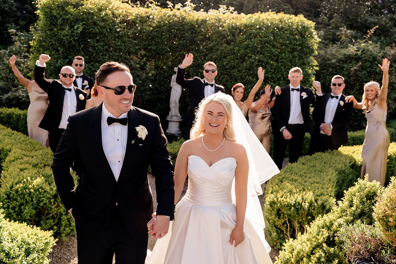 Wedding party celebrating in a lush garden setting with a couple in white dress and black tuxedo leading the group.