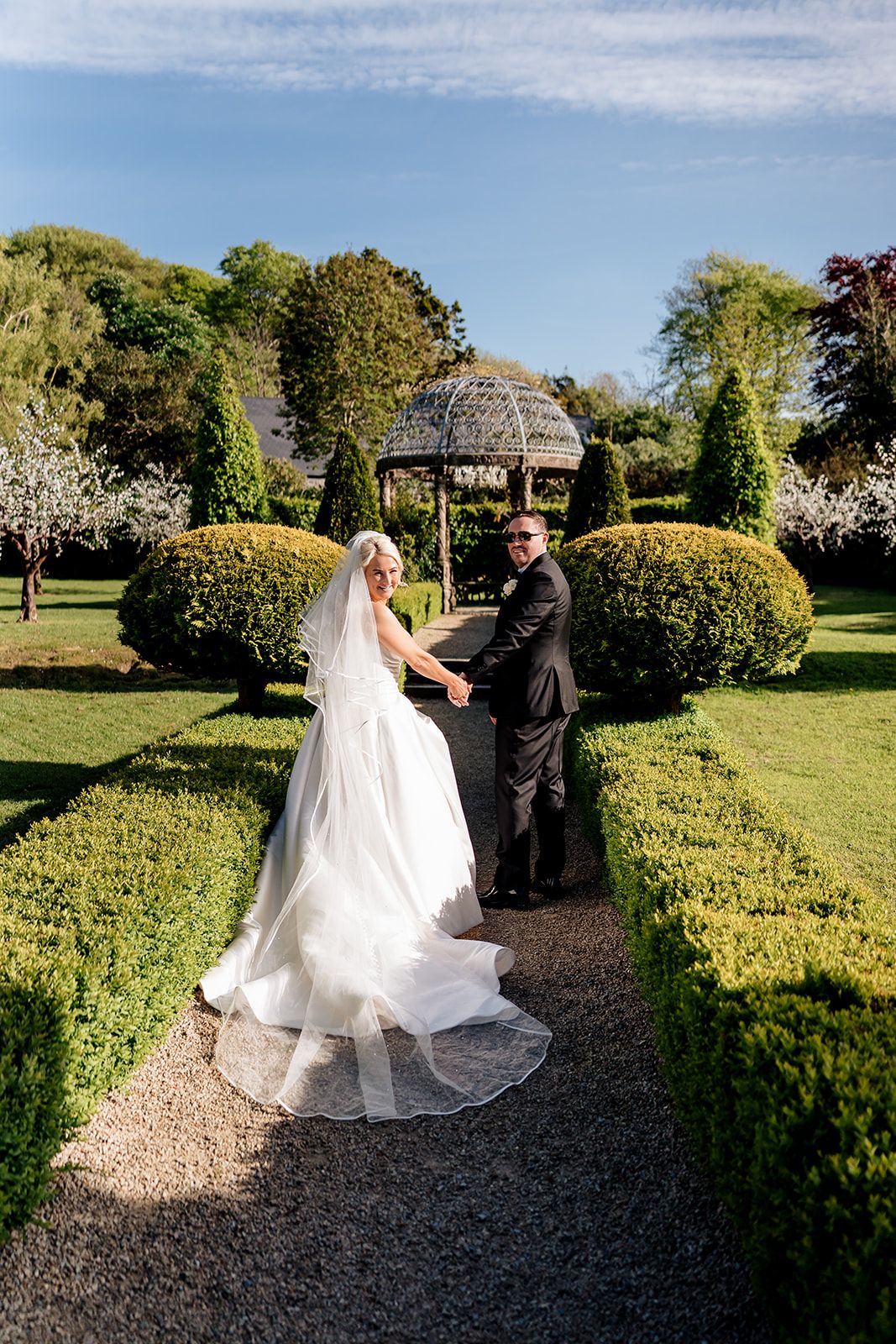 A couple stands in a formal garden with manicured hedges and a gazebo on a sunny spring day.