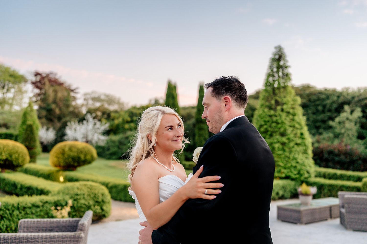 A wedding couple shares a joyful moment together in a manicured garden with topiary and landscaping at sunset.