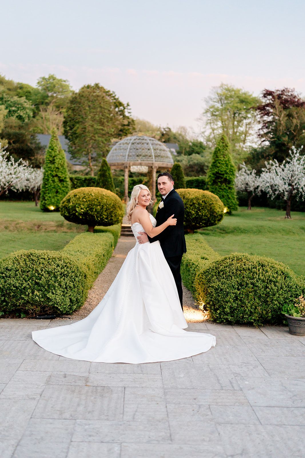 A married couple poses in front of a garden gazebo with manicured hedges and spring blossoms in the background.