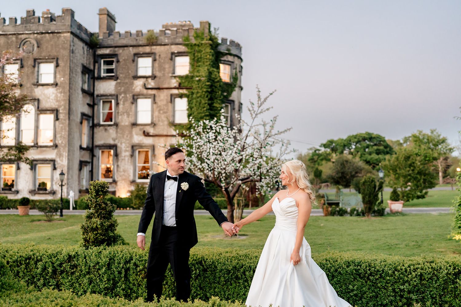 Elegant wedding couple poses on manicured lawn beside historic stone castle ruin with ivy covered walls.
