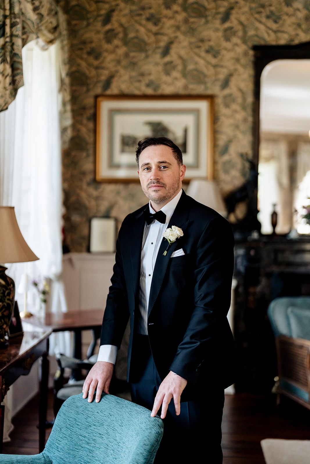 A formal wedding photo showing a groom in a classic black tuxedo and white boutonniere standing in an elegant room.