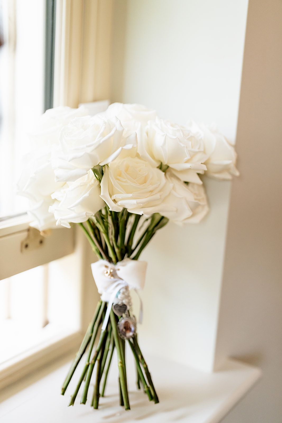 Simple elegant white rose bouquet with green stems tied in ribbon sits on windowsill in soft natural light.