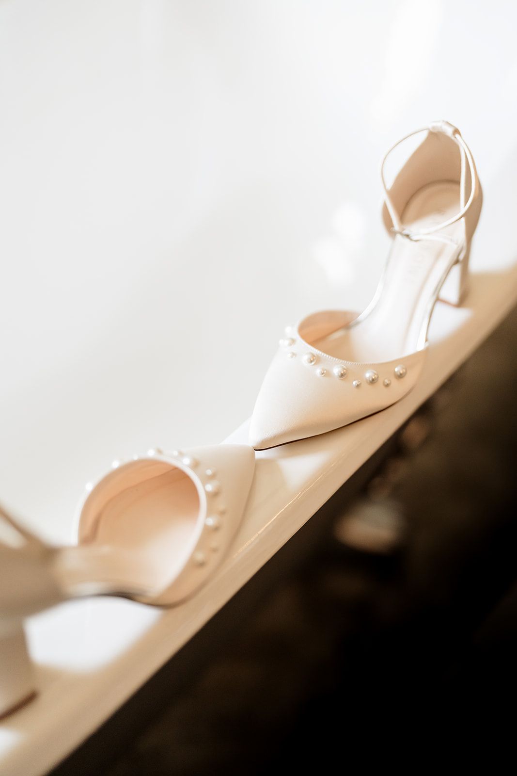 Elegant white bridal heels with pearl details sit artistically on a shadowy surface.