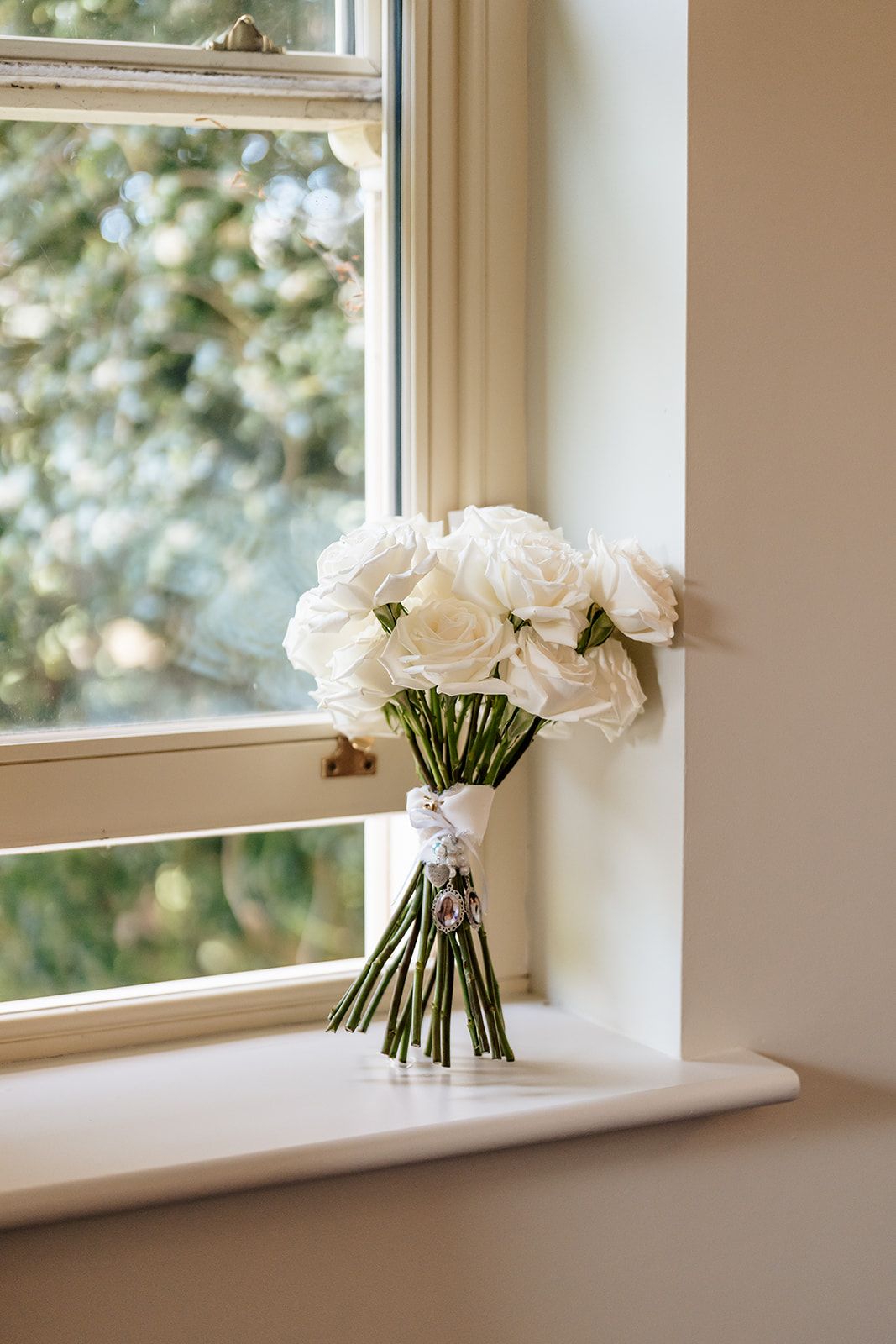 A delicate bouquet of white roses tied with ribbon sits on a bright windowsill, with soft natural light filtering through.