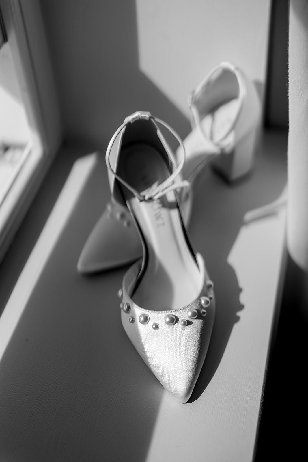 A black and white photograph of an elegant white bridal shoe with rhinestone details resting near a window.