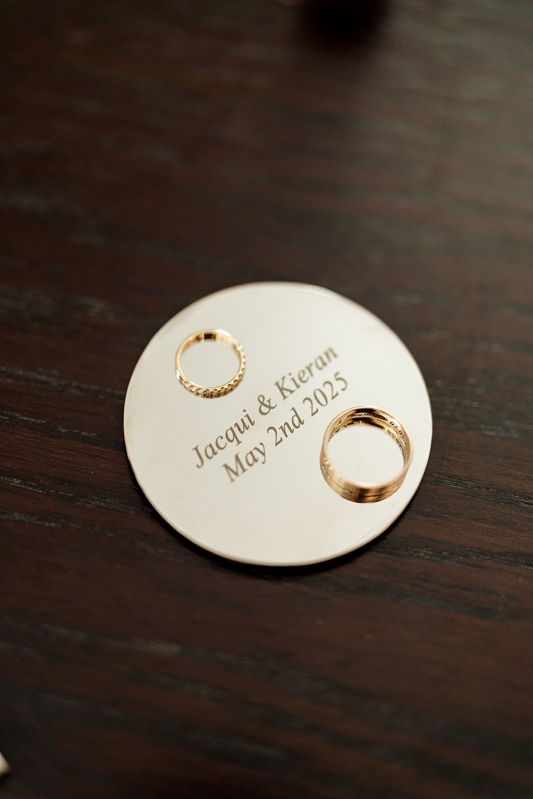 Gold wedding rings and names engraved on a white circular surface against dark wooden background.