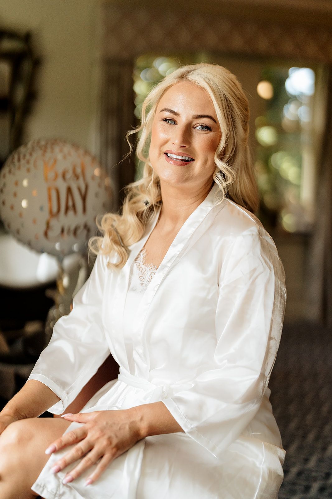 Woman in white silk robe sitting on couch with Best Day Ever balloon decoration in background.