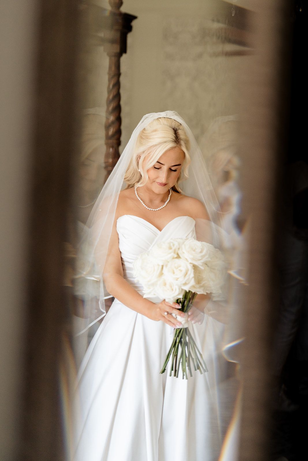 Bride in elegant white strapless gown holds a beautiful white rose bouquet while gazing down in a soft-focus setting.