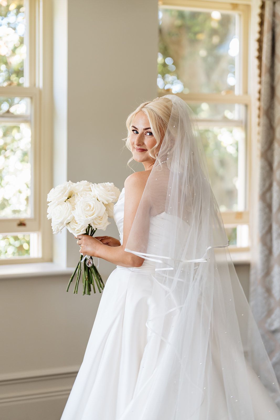 A bride in a classic white wedding dress holds a bouquet of white roses while standing in a sunlit room.