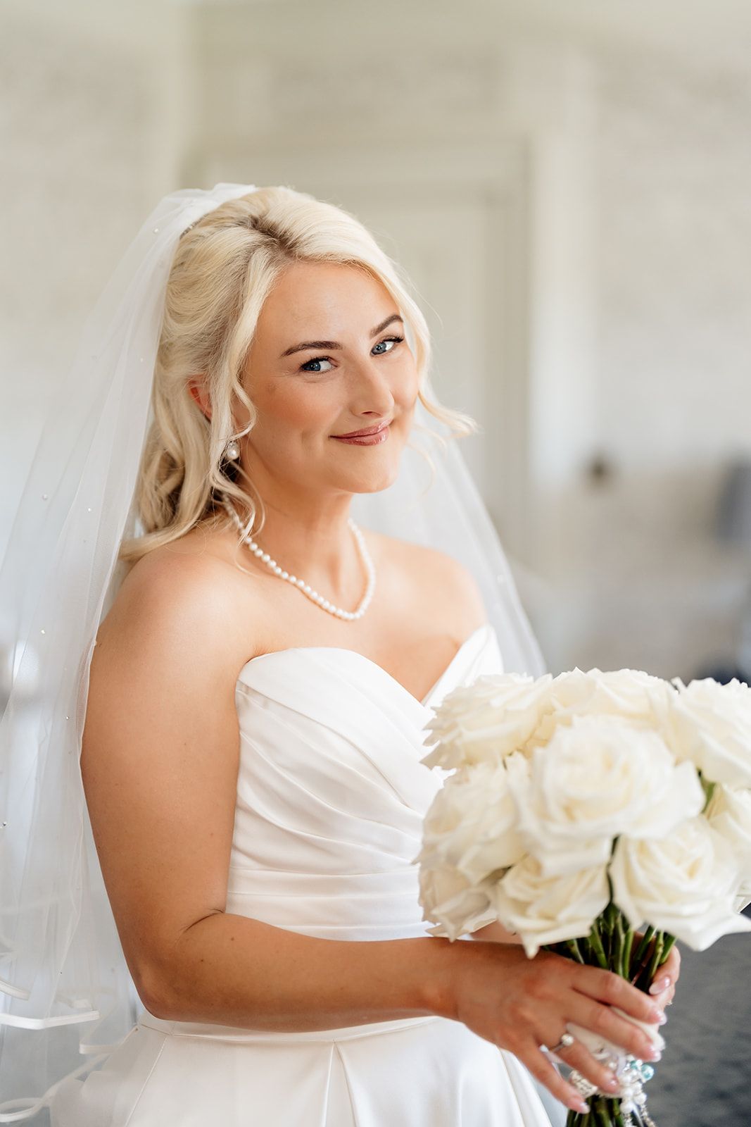 Beautiful bride in white strapless dress holds elegant bouquet of white roses while wearing pearl jewelry and veil.
