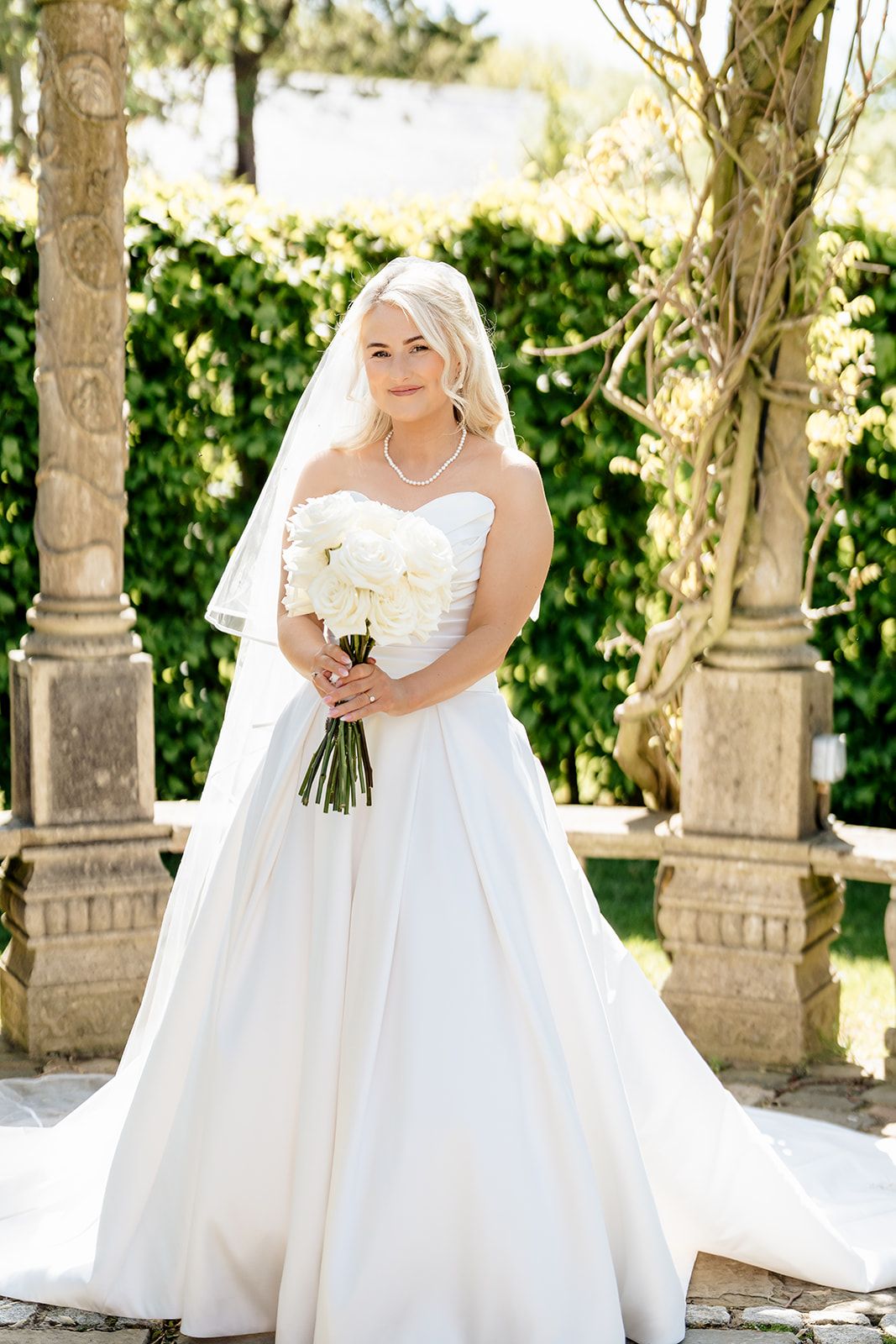 Bride in strapless ballgown holding bouquet poses between stone columns in garden setting.