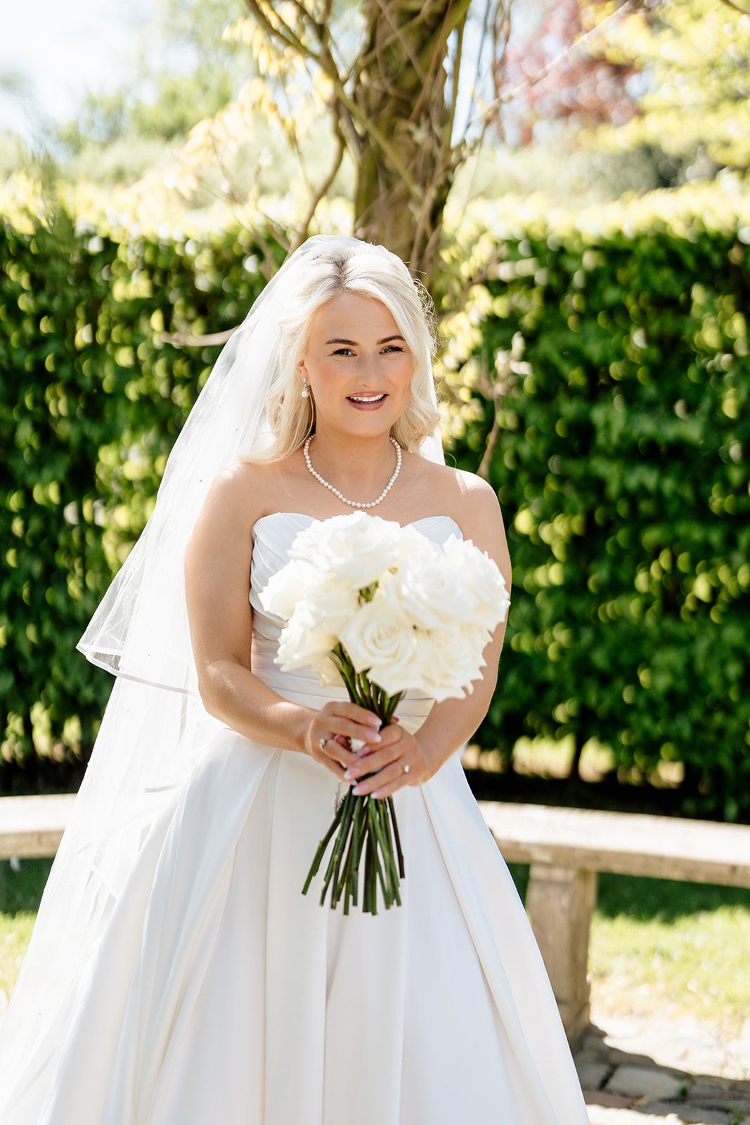 Beautiful bride in white strapless gown holds elegant white peony bouquet outdoors on sunny wedding day.