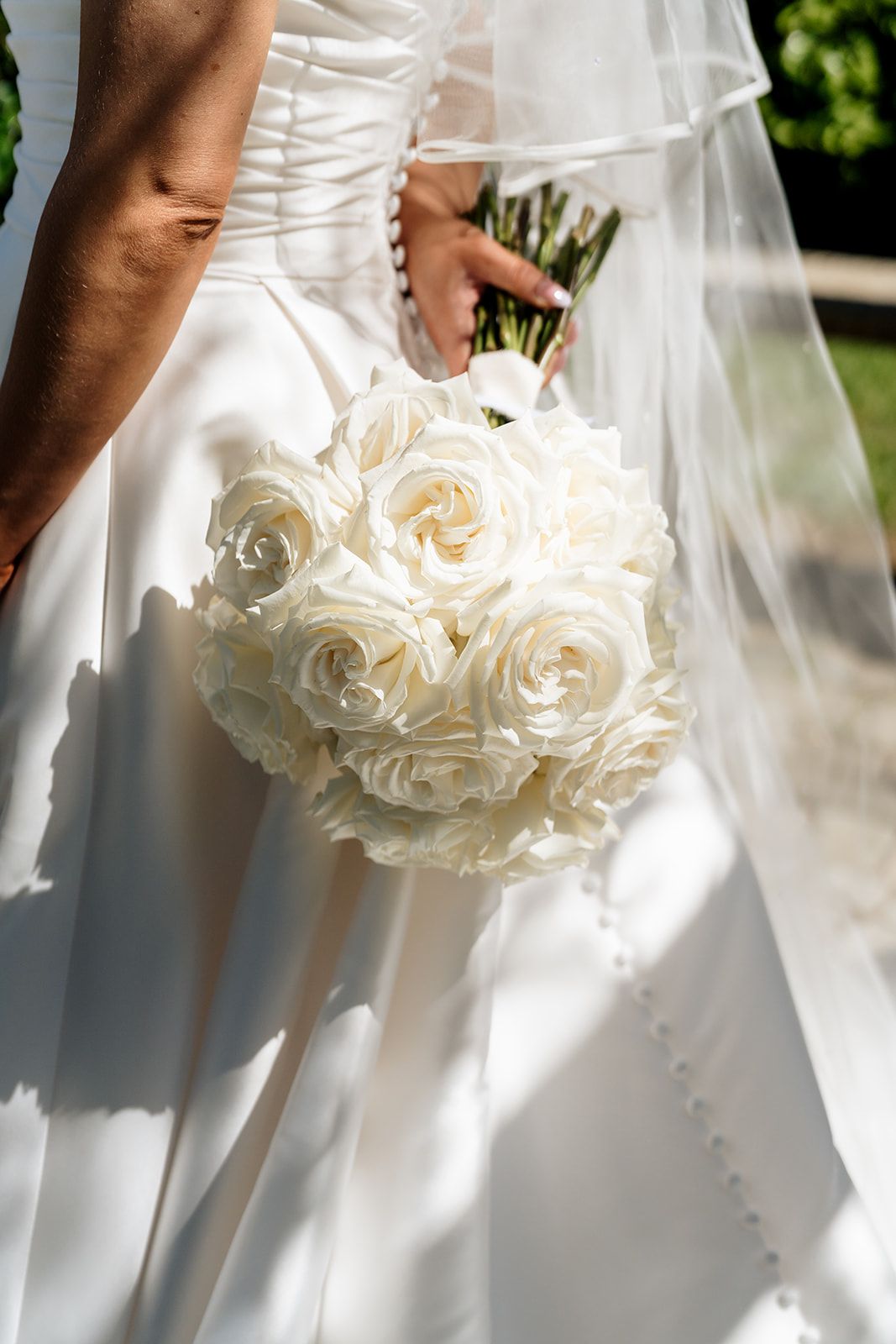Close-up of an elegant white rose bridal bouquet against a wedding dress with sunlight casting soft shadows.
