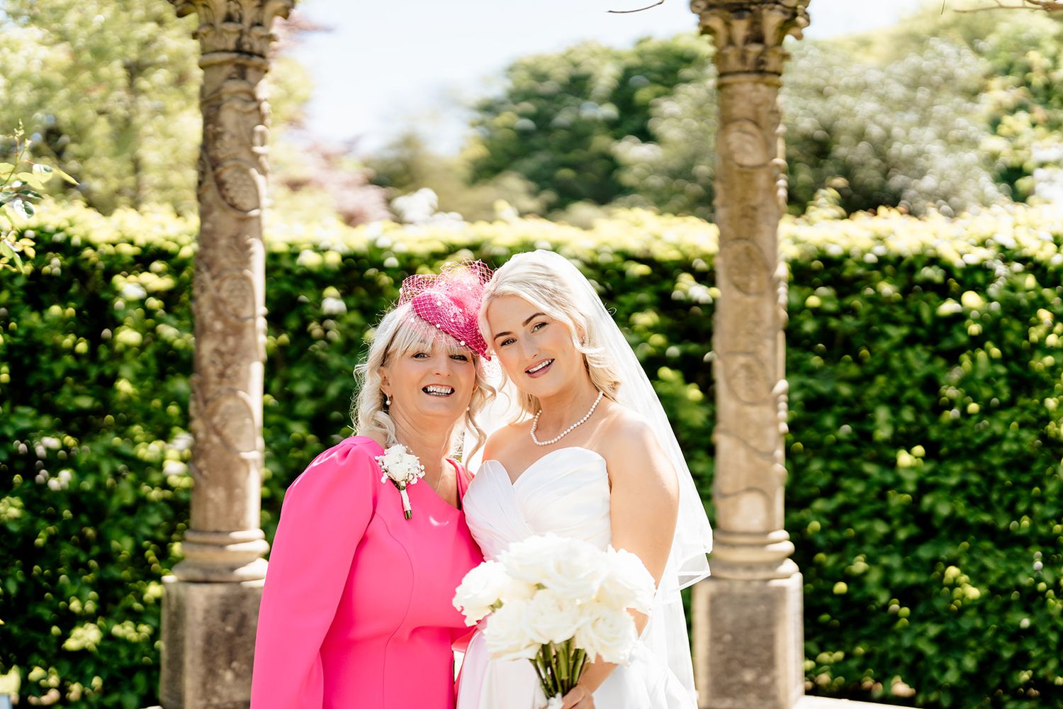 Wedding photo taken in a garden with stone pillars and lush hedges in the background.