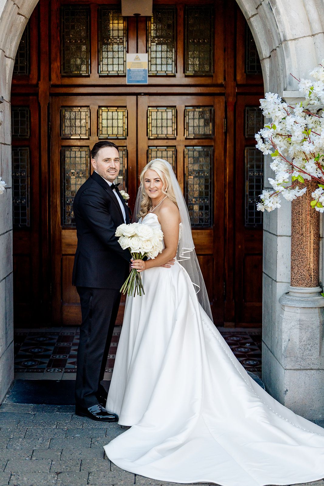 A newlywed couple poses in front of an ornate wooden door with stained glass panels and white spring blossoms.