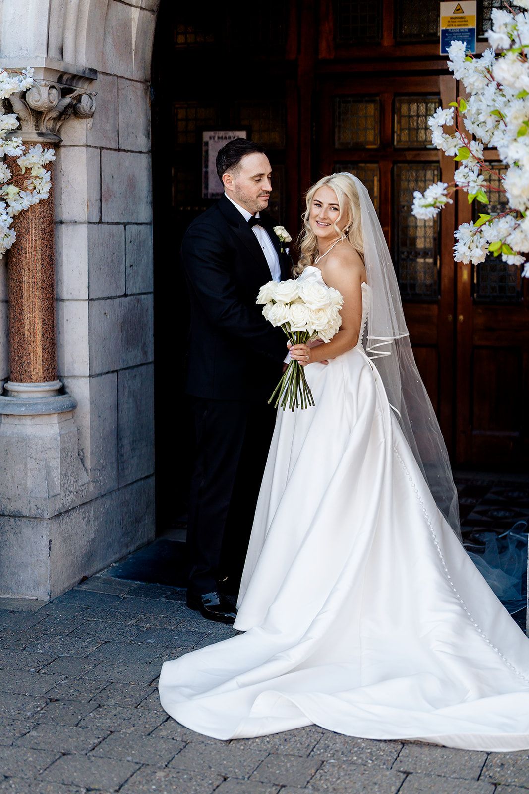 A bride in a white wedding gown with a long train and veil holds a bouquet while posing with groom in tuxedo by stone archway.