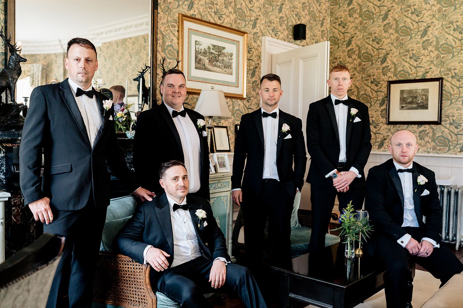 A group of groomsmen in black tuxedos pose together in an elegant room with patterned wallpaper.