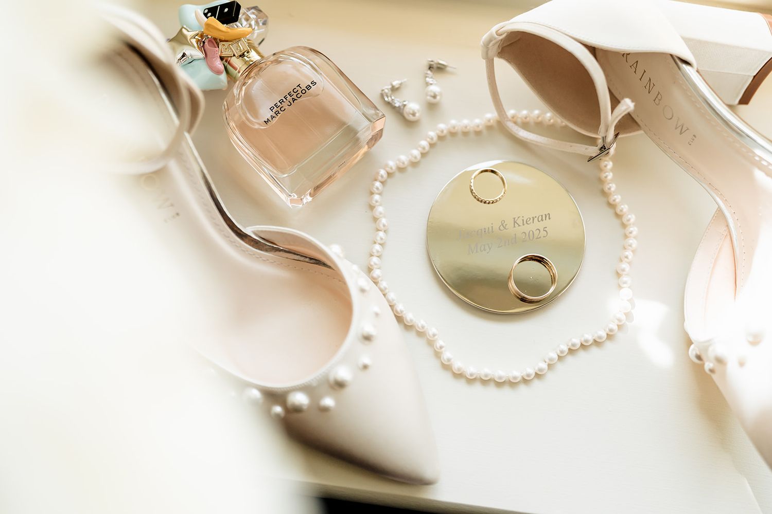 Wedding accessories including pearl necklaces, gold rings, heels, and a perfume bottle arranged on a light surface.