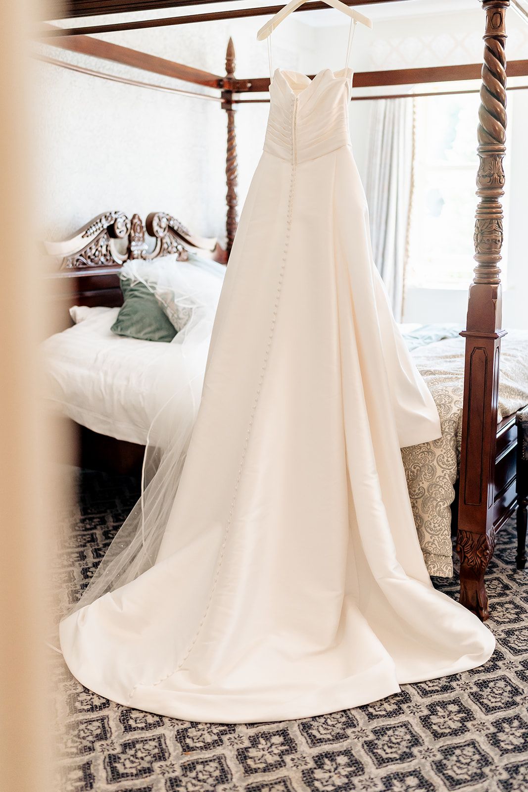 An elegant white wedding dress with a long train hangs from a wooden four-poster bed over a patterned carpet.