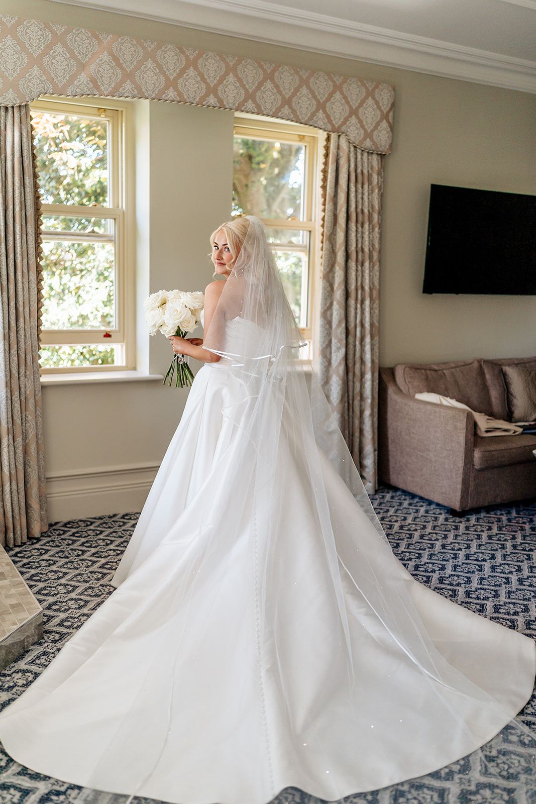 A flowing white wedding gown with cathedral train and veil stands elegantly in front of sunlit windows in a formal room.