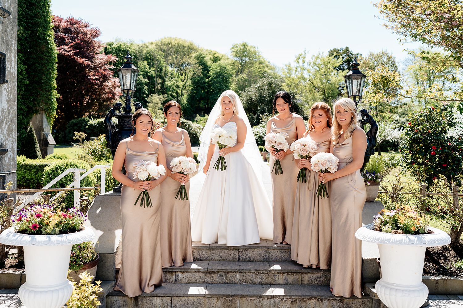 A wedding party in beige bridesmaid dresses and white wedding gown pose on stone steps in a sunny garden setting.