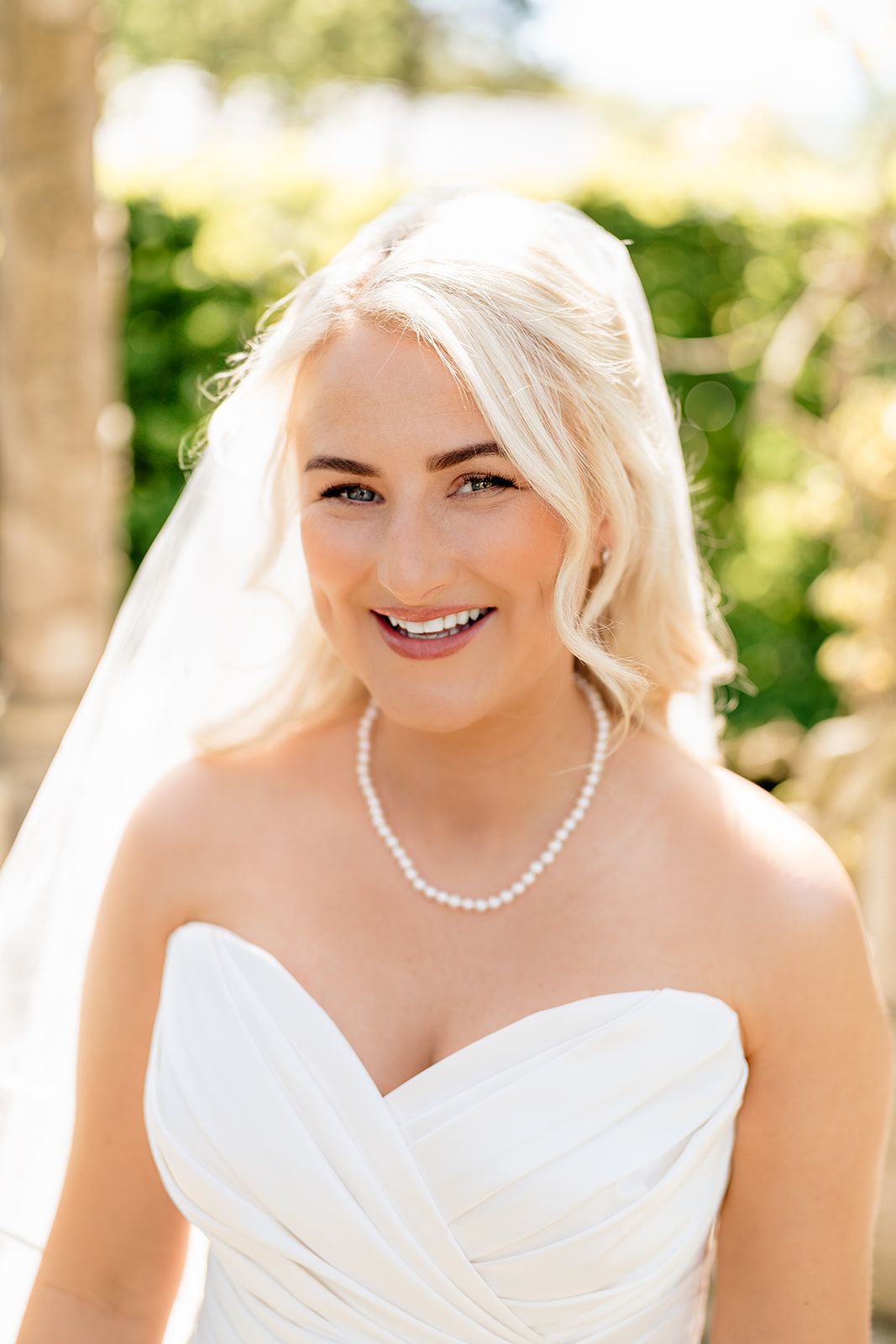 A bride wearing a white strapless wedding dress and pearl necklace smiles outdoors on her wedding day.