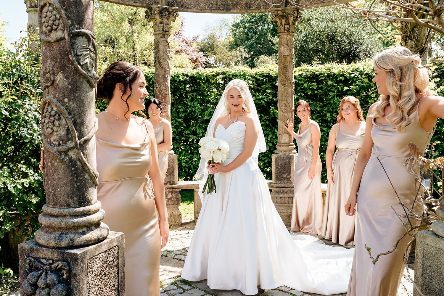Bridal party gathers under an elegant stone pergola during an outdoor wedding ceremony.