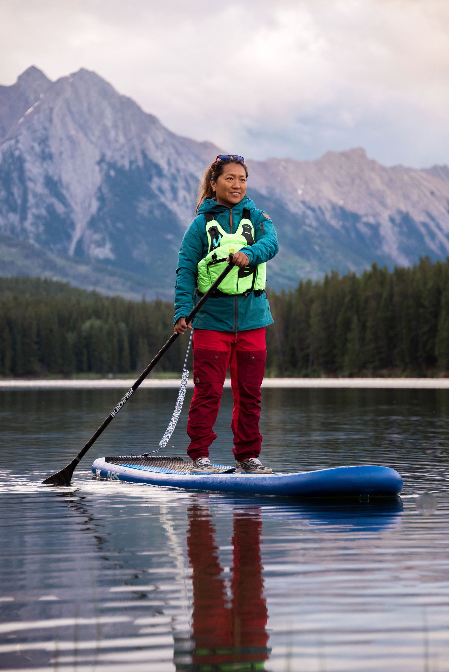 Person paddleboarding on calm mountain lake with stunning rocky peaks and forest in background at dusk.
