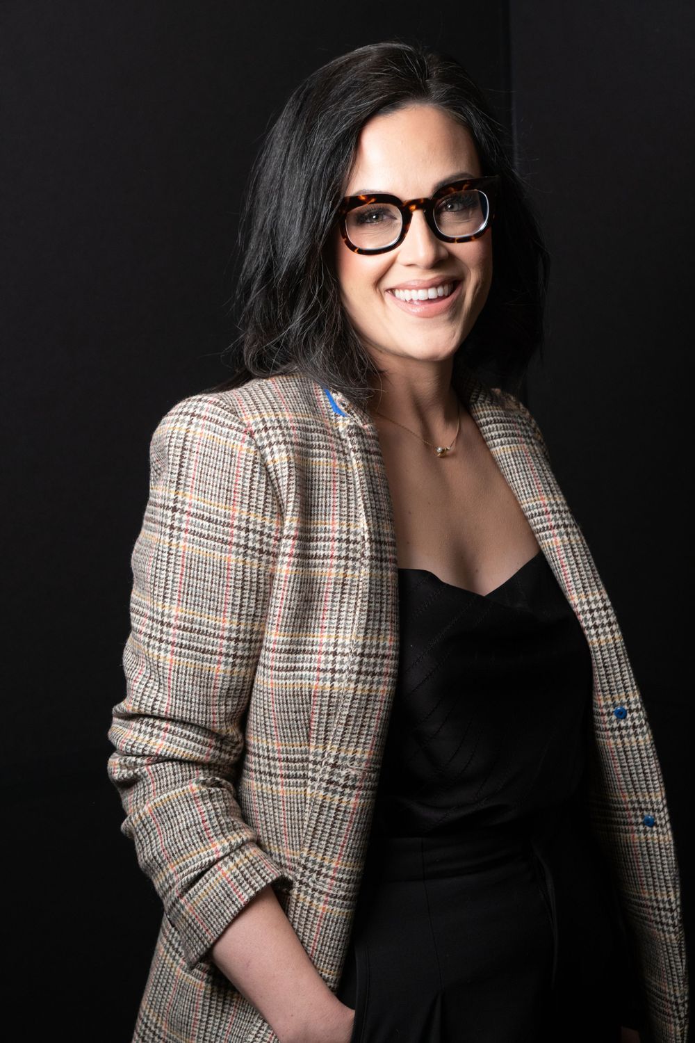 Professional headshot featuring plaid blazer, black top and stylish glasses against dark background.