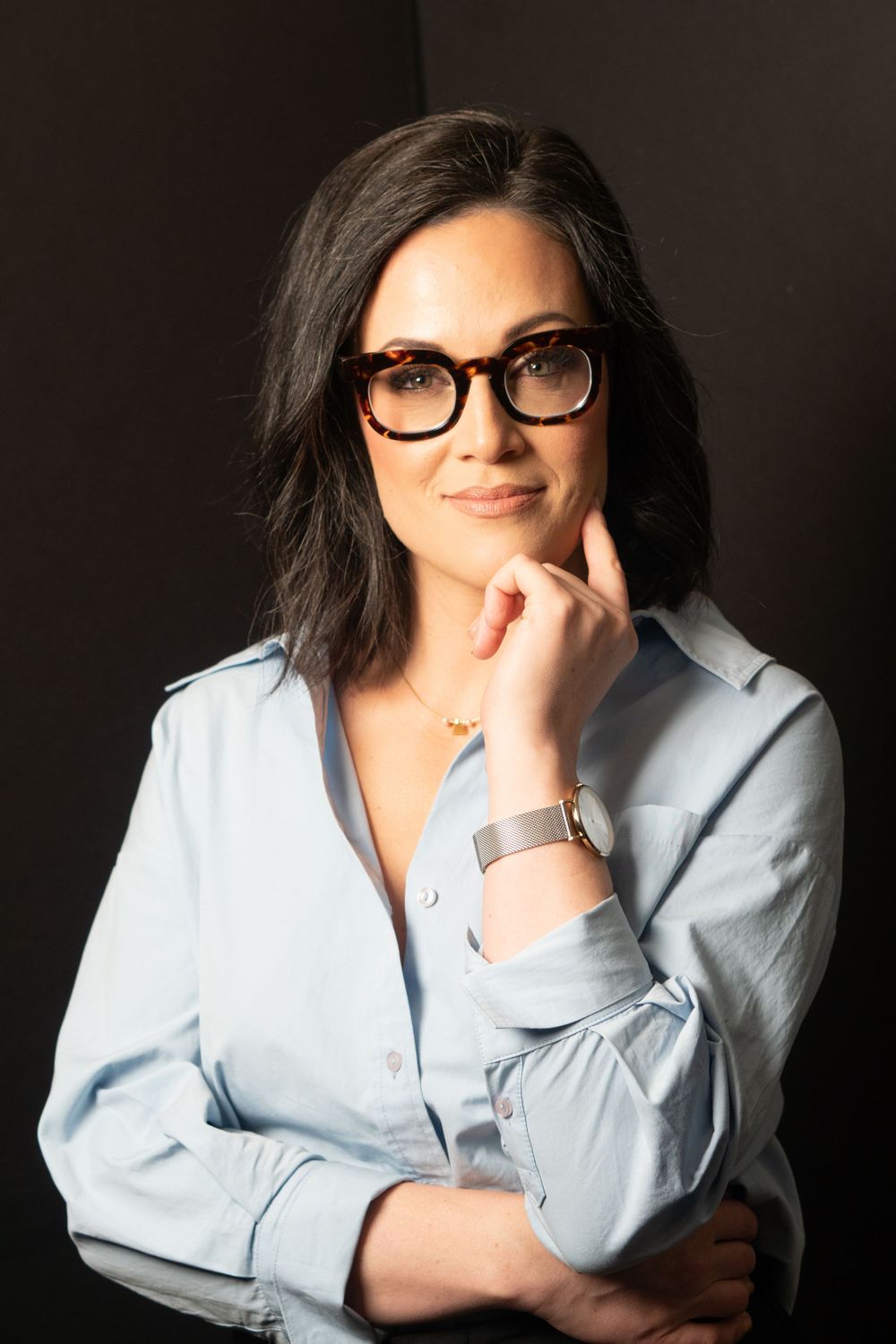 Professional portrait with thoughtful pose wearing light blue shirt and glasses against dark backdrop.