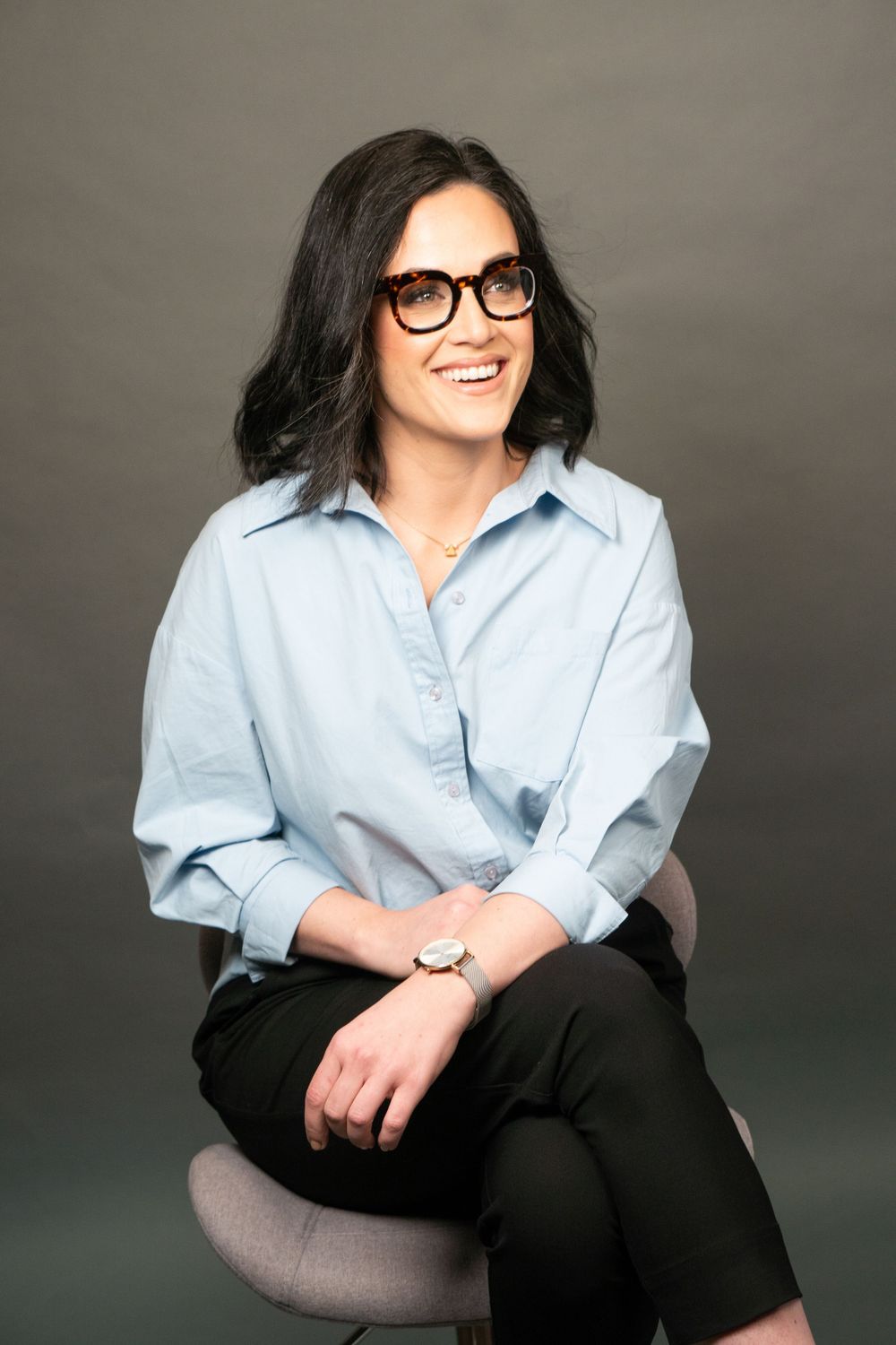 Business portrait of person in light blue button-down shirt seated on gray chair, smiling and looking out in the distance.