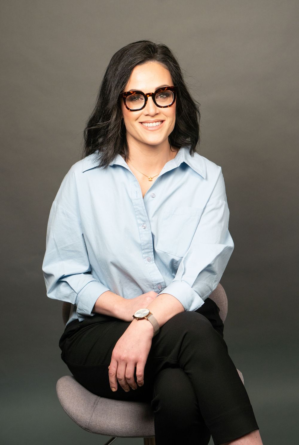 Professional studio photo with light blue shirt and black pants seated on gray chair.