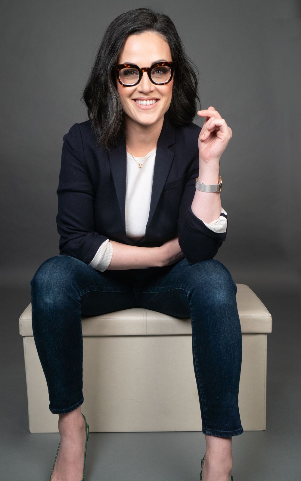 Studio portrait showing relaxed pose on light colored cube wearing business casual attire and glasses.