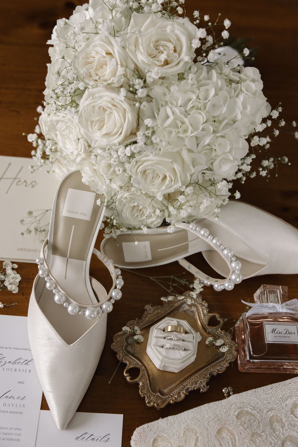 Elegant white wedding accessories with roses, bridal shoes, perfume bottle and pearl details on a wooden surface.