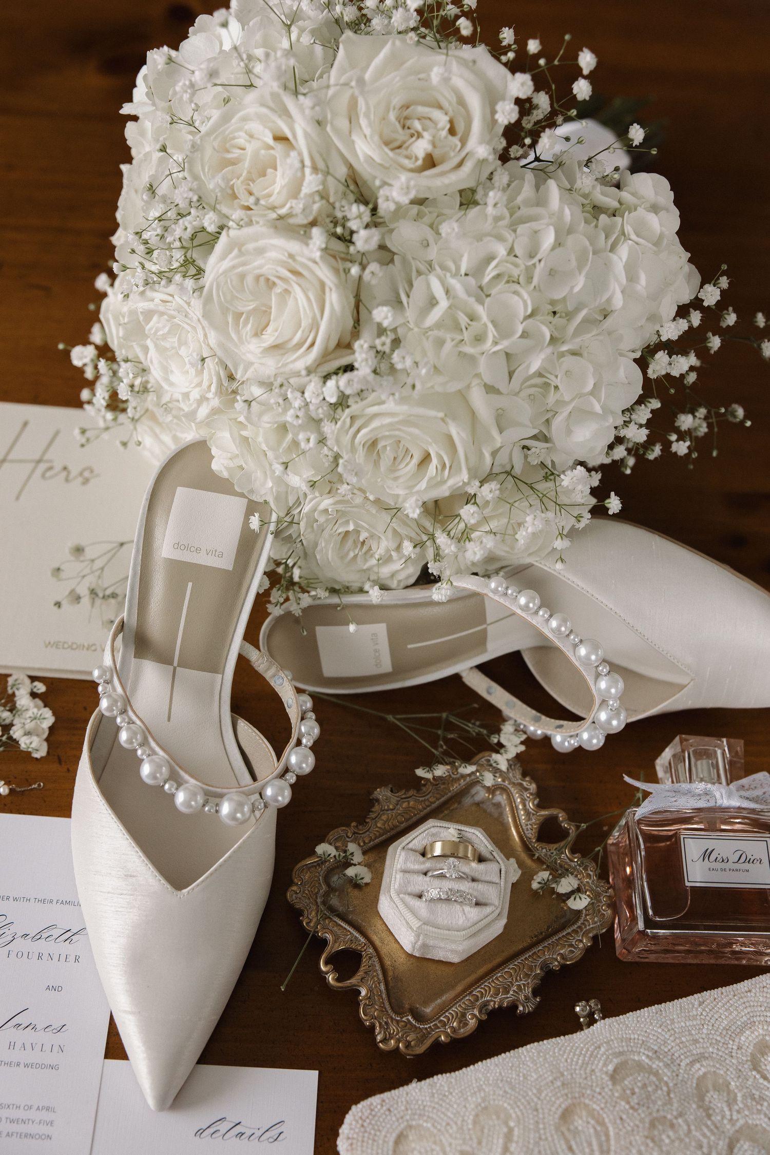 Elegant white wedding accessories with roses, bridal shoes, perfume bottle and pearl details on a wooden surface.