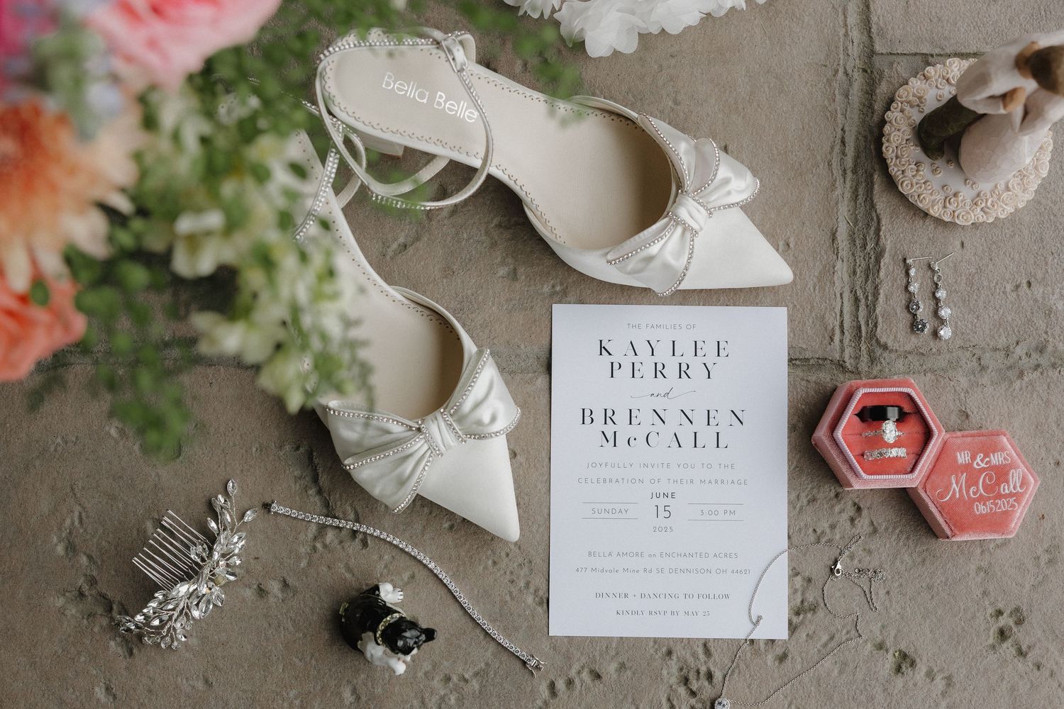Wedding details flatlay with white bridal shoes, invitation card, and floral accents on concrete surface.