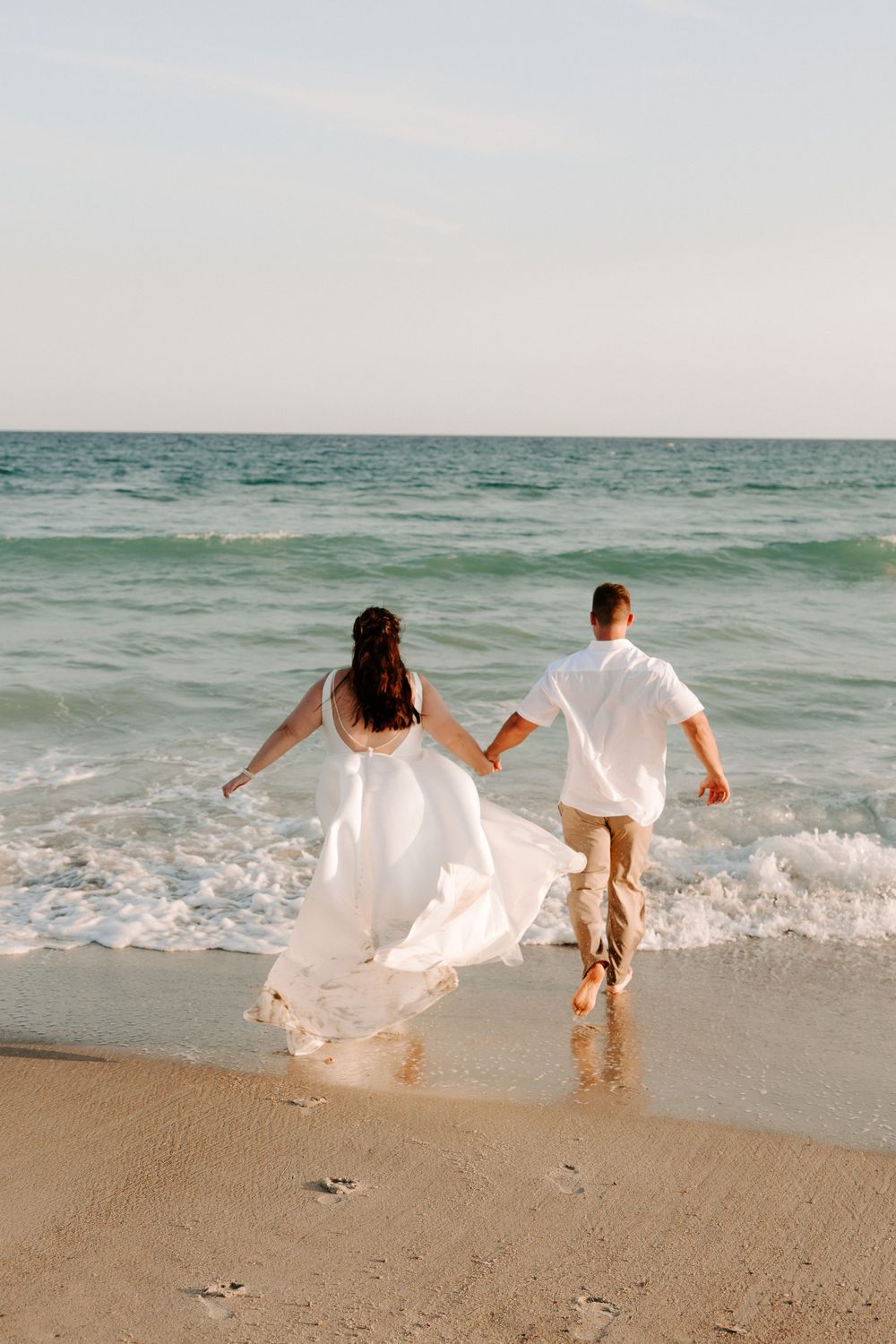 Couple running through ocean waves on beach in wedding attire.