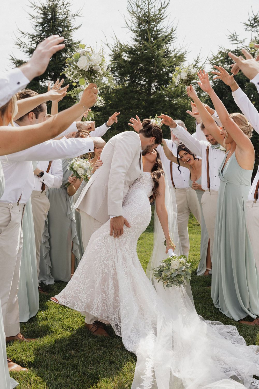 Wedding party celebrates with raised hands as couple shares romantic moment on grassy lawn with evergreen trees.