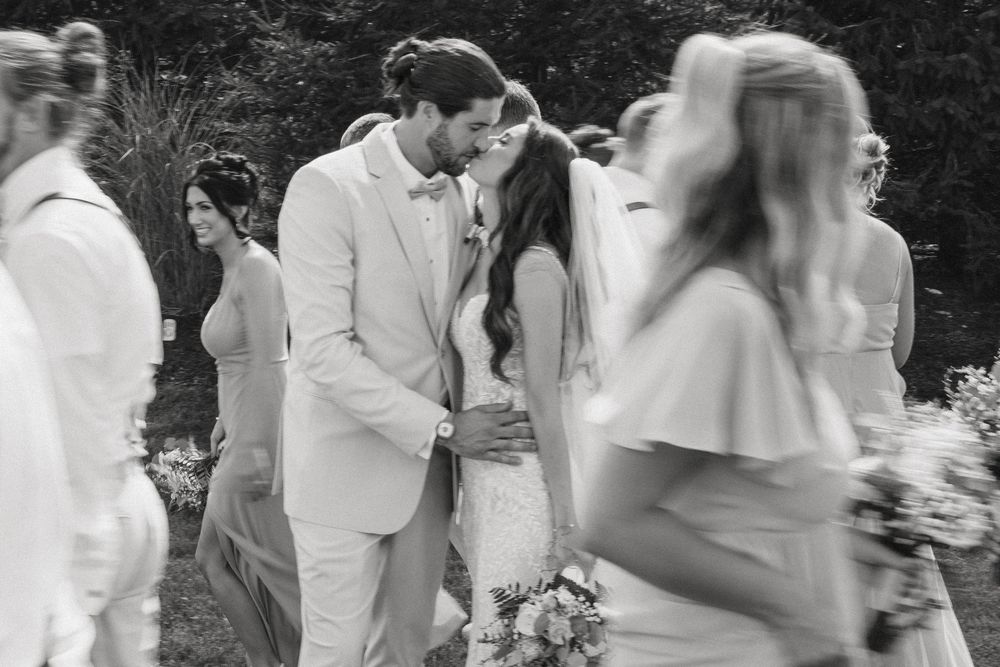 Black and white photo of a couple sharing a tender moment at an outdoor wedding ceremony surrounded by guests in white.