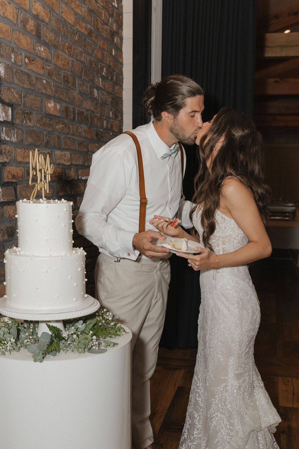 Newlywed couple shares a slice of white wedding cake at rustic brick venue with baby's breath floral accents.