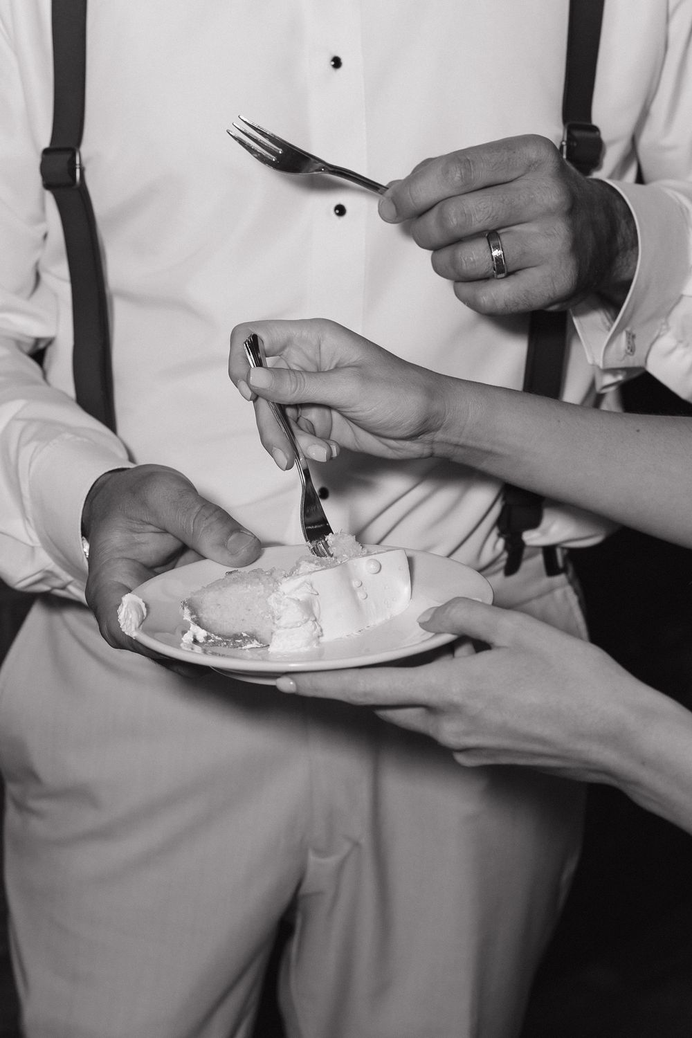 Black and white photo showing hands holding plates and utensils while eating at a dining table.